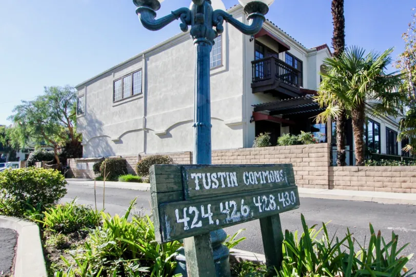 A sunny day by the Tustin Commons sign in front of a light pole with trees and houses behind the light pole