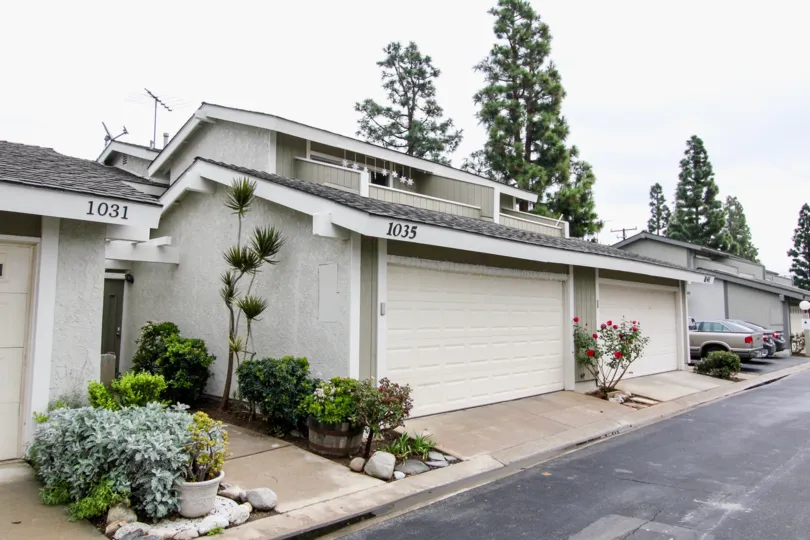 An overcast day by a block of Tustin Pines houses with three cars parked in the driveway at the end and tall trees behind the houses