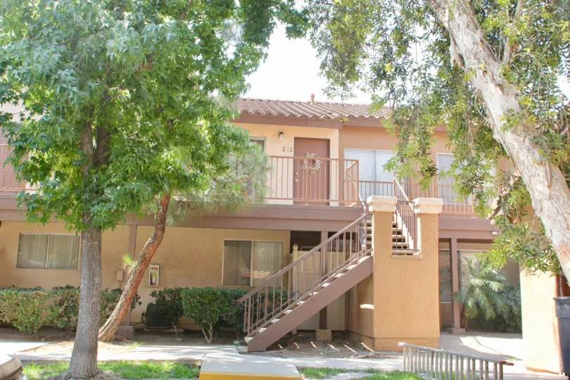 there is a stair case in front of house to reach first floor and house is surrounded by trees and grasses and it is sunny day
