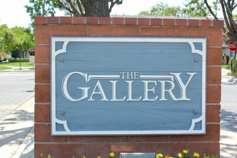 Entrance sign made of brick and blue background saying The Gallery with flowers planted and gardens in the distance