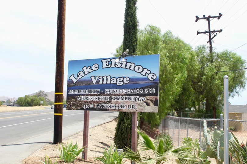 Entrance sign with sandy area with palm trees and large trees near power lines entrance