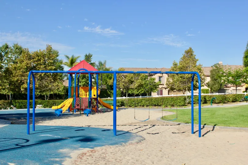 Fenced playground area with sand surface and colorful structures with multiple swings and slides