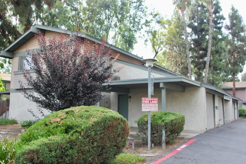 Tan, two-story apartment building with four single car garages adjacent to the parking lot. Small green bushes in front of the building, and pine trees behind the building
