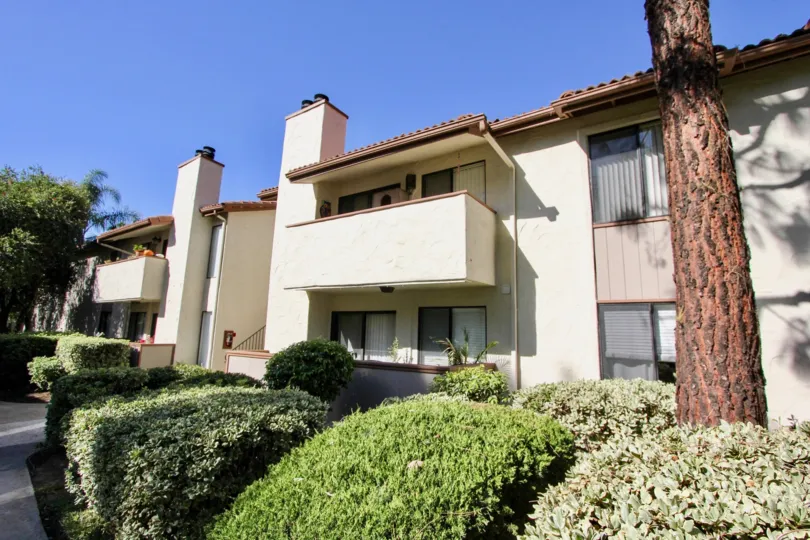 An tan, 2 story apartment complex with a balcony on a sunny day with a tree and shrubs.