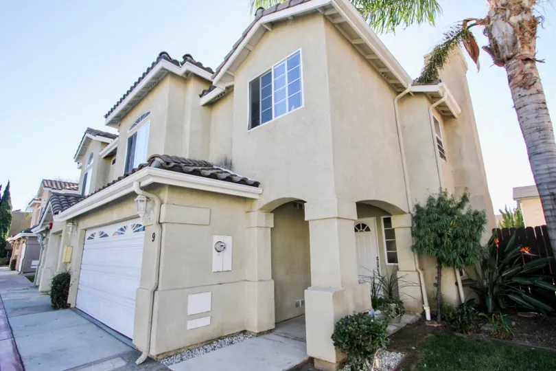 Moss Villas, Chula Vista, California. A two-story house with a cream-colored exterior and a white garage sits in a neighborhood of similar houses. The sky is bright blue. There is a palm tree in the yard.