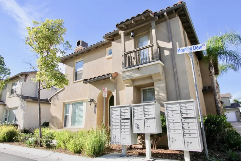 2 story house with small balcony, mailboxes, trees, and small bushes on a sunny day.