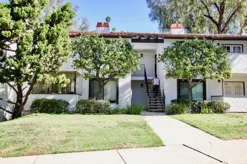 A white complex with 3 trees out front and some shrubbery. There is a man walking up the stairs. Located in Collwood Meadows of College Area, California