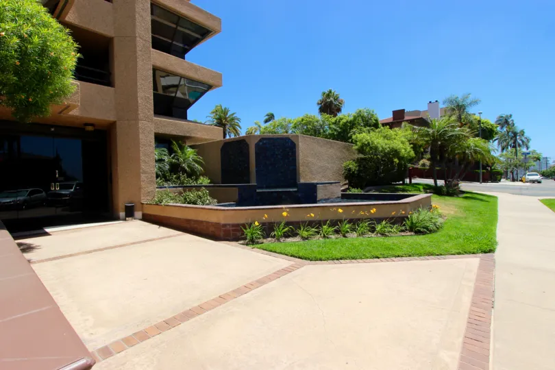 A sunny day with a brown house is decorated with greenery, walkway leading to the house has a small lake