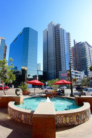 A sunny and brilliant day at the Village walk with a cheerful, rejuvenating fountain and small shops against a a backdrop of a panoramic view of massive high rise buildings