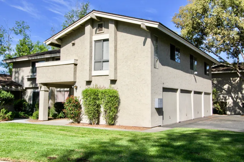 Tan stucco house with a four car garadge nice green lawn and a couple bushes next to the balcony