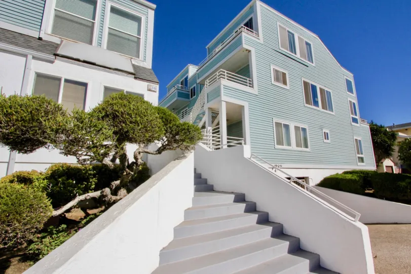 A staircase going to a light-blue apartment building and some bushes in the side during a sunny day in Coastwalk Townhomes, Imperial Beach, CA