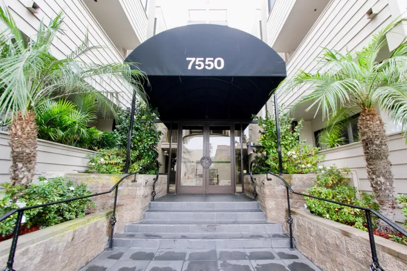 A regular hotel entrance with a few stairs, a rain cover and vegetation all around it.