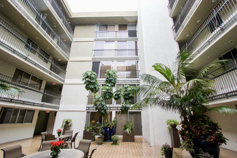 The patio at Devonshire, La Jolla, CA, with some palm trees, tables and chairs, and a view of the apartments