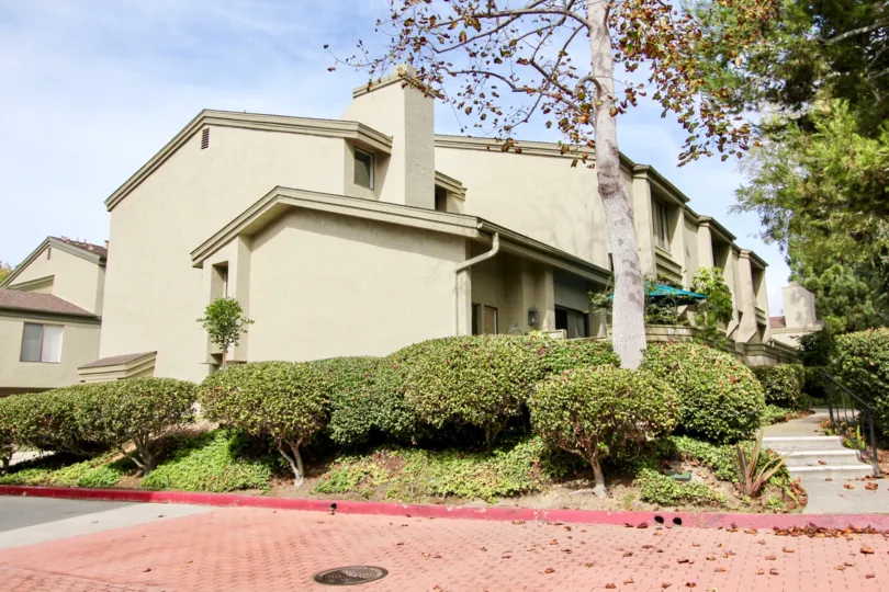 A sand-color house with some bushes and trees in front and a pathway on the side during a sunny day in Eastbluff, La Jolla, CA