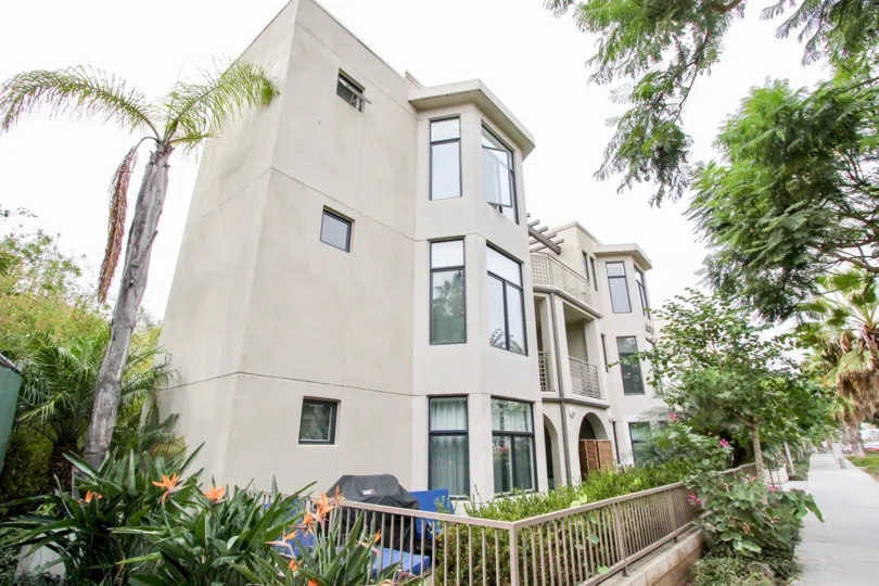 A whilte three story apartment complet with big windows and lots of plants and trees both in the garden and on the street.