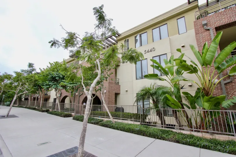 A yellow three story apartment complex with a balcony. on the pavement and outside of the apartment are a lot of palms and other trees.