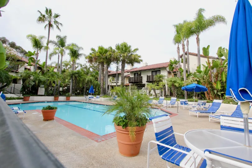 An empty swimming pool on a cloudy day with chairs, tables, plants and palm trees around apartments