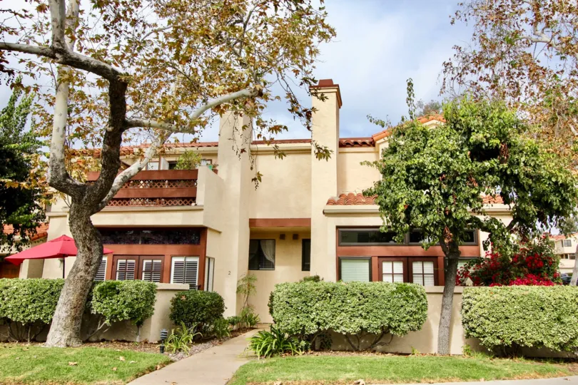 A coffee and cream stuccoed 2 story duplex with clay tiled roof and 2 chimneys hides patios behind a wall, bushes and trees