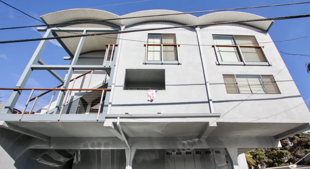 Grey building in the community of Mission shores. This building is located in Mission Beach, California. Blue sky in the background of this upward facing picture of this two story building. Pictured are three windows and a flight of stairs.