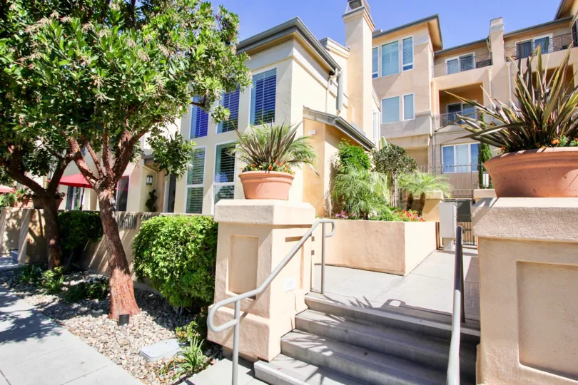 The entrance to an apartment building with some plants in pots, bushes and trees during a sunny day in Wellington Square, Mission Hills, CA
