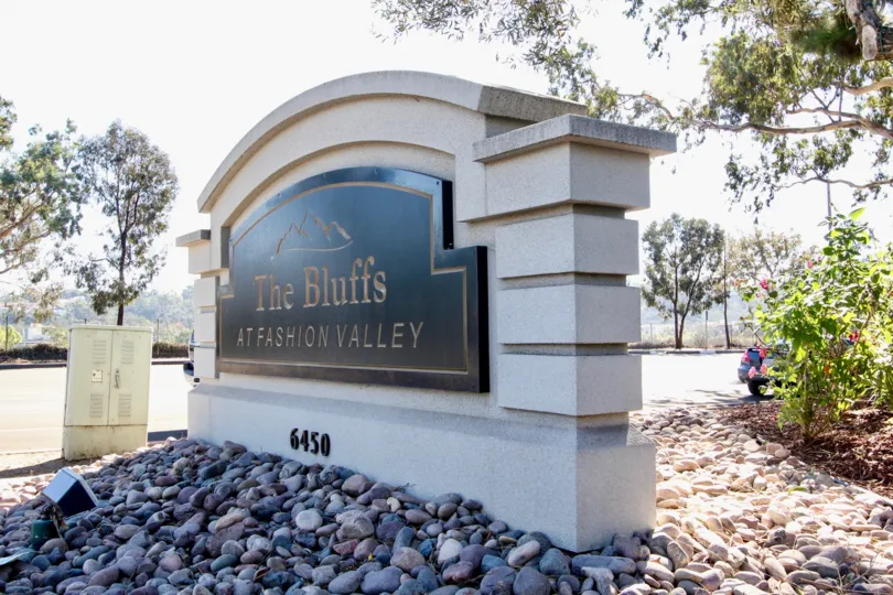 Sign of "The Bluffs at Fashion Valley" in Mission Valley, California during the daytime. Sign is on top of stones with trees in background.