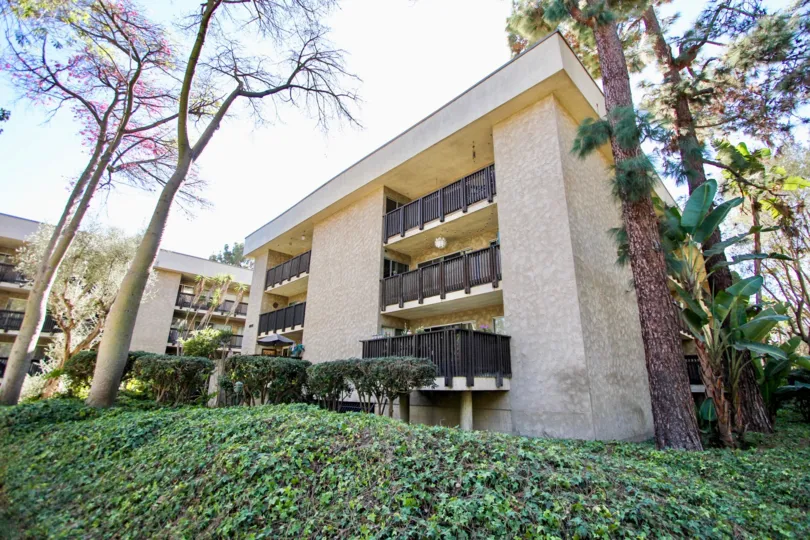 Outside view of The Bluffs building in Mission Valley, CA. Shrubs and trees surround the 3-story building. Another building in the background.