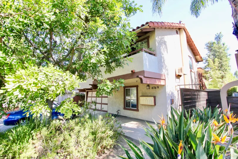 A two-story unit with a garage and balcony is surrounded by greenery and abundant sunshine