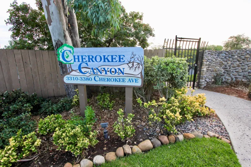 A welcoming sign for Cherokee Canyon.  Nice lanscaping with shrubs bordered by rocks.  A metal gate with a stone fence on one side and a wooden fence on the other insures privacy.