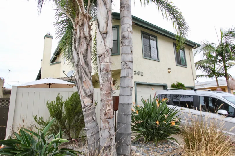 This is a single family home in Park Villas Community in North Park, California. It has a light painted facade with green windows and the property number is 3412. It also has a palm tree and a driveway in the front yard.