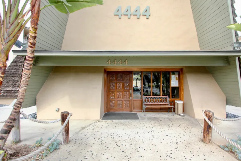 The front walkway of Point Loma Villas with palm trees, a bench, garbage can, windows, wooden door and a fence of rope and logs.