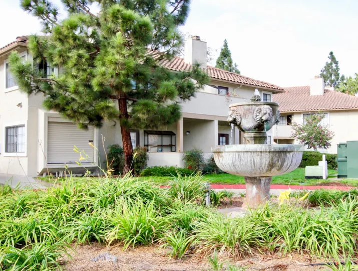 A view showing the exterior of the Vista Way Village with a central fountain, landscaping, and tile-roofed apartment units.