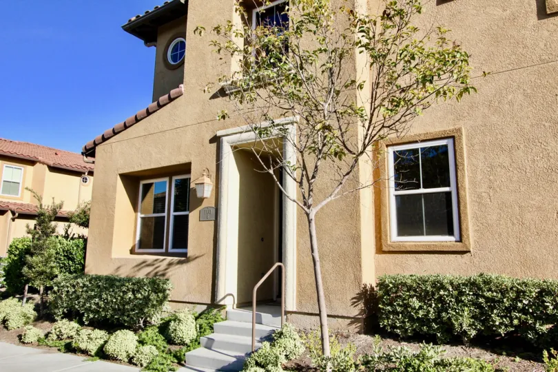 Beige front of house with green shrubs and a young tree, terracotta roof