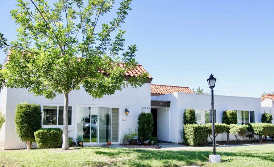 A white house with red roofing with a some bushes, a tree and a lamp in front during a sunny day in Haciendas, Rancho Bernardo, CA