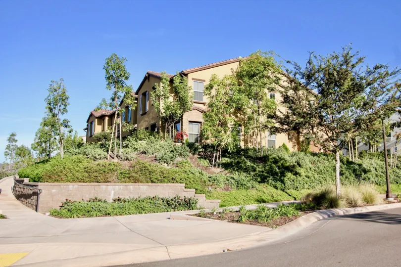 A yellow house up a hill with some trees growing around it and a street in front during a sunny day in Mandolin, Rancho Bernardo, CA