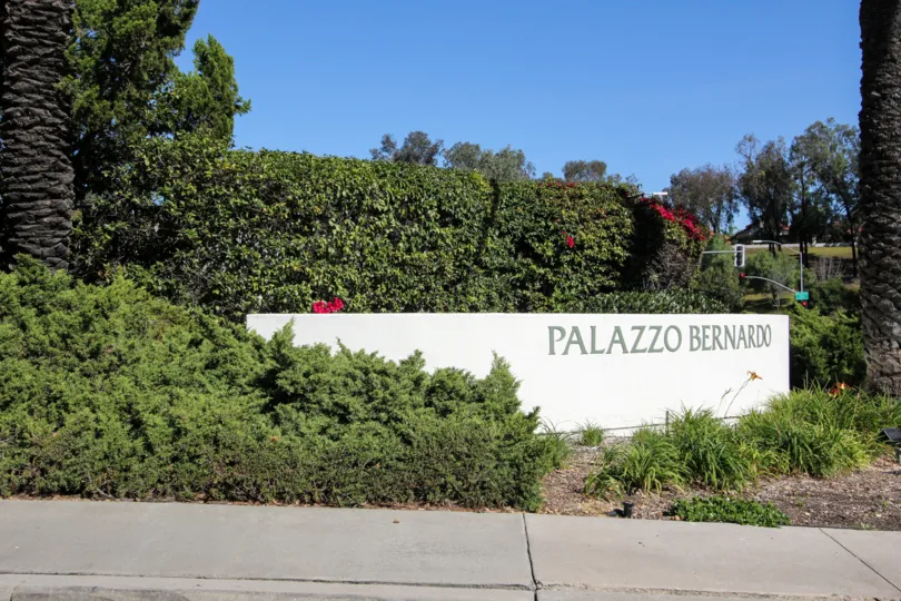 Sidewalk and lush greenery surrounding signage in front of the Palazzo Bernardo in California community.