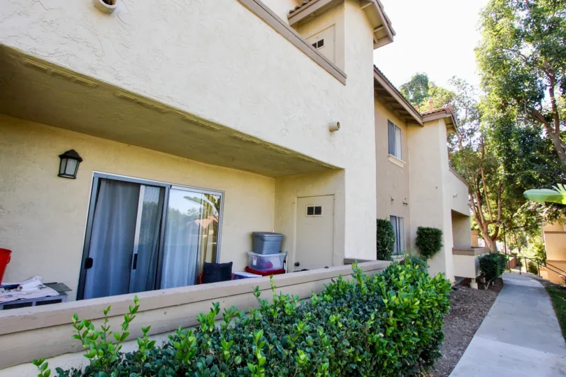 A sand-color house with some bushes and trees surounding it and a pathway to the side during a sunny day in Mission Park, San Marcos, CA
