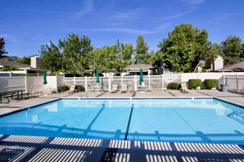 A large swimming pool with some recliner chairs, a white fence and trees and houses in the back during a sunny day in Shadowridge Greens, Vista, CA
