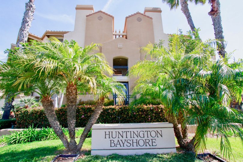 A HOUSE WITH A BOARD OF SIGN IT REPRESENTS HUNTINGTON BAYSHORE AND SOME TREES ALSO BOTH SIDE OF THE BOARD AND A CUTE HOUSE ALSO AT THE BACKSIDE OF THE BOARD SIGN