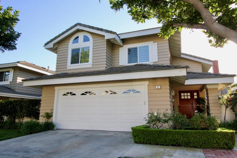 A sunny day in Ridge Garden Homes with a house with a garage for two cars and brown front door and tall tree next to it.