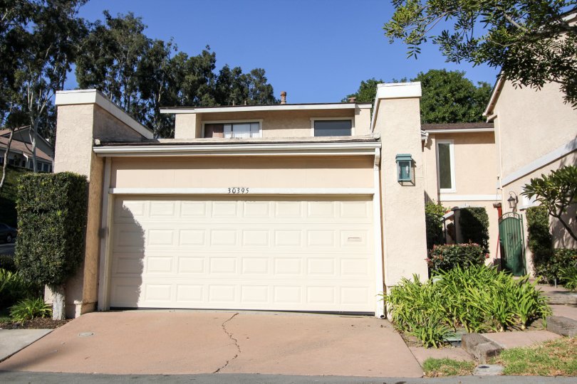 A sunny day by a El Niguel Terrace house with trees behind it and other greenery on each side of the walkway to the front door and the garage door, along with a crack in the driveway concrete