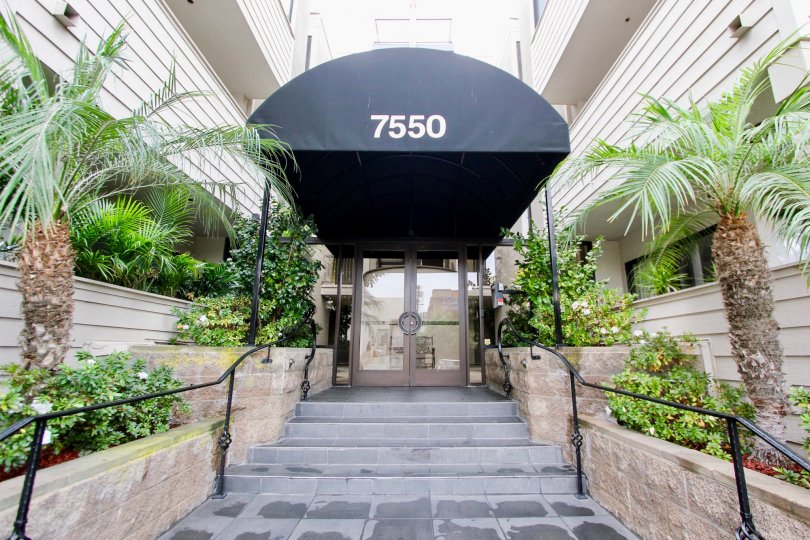 A regular hotel entrance with a few stairs, a rain cover and vegetation all around it.